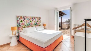 A bedroom with a bed, a nightstand, a lamp, a colorful floral headboard, and a view of the outside. - Villa Chez Piè: Panoramic Views, Pool & Concierge (Massa Lubrense)