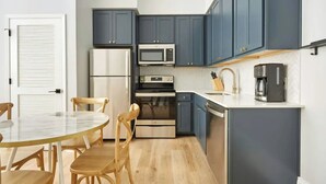 Eine kompakte Küche mit dunkelblauen Schränken, einem Edelstahl-Kühlschrank, einer Mikrowelle, einem Backofen und einer Kaffeemaschine. Ein Holztisch mit Stühlen steht daneben.