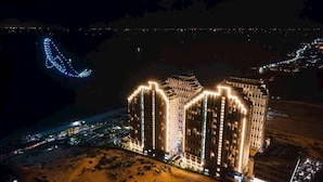 Un hôtel de grande hauteur, composé de plusieurs étages et illuminé par des éclairages extérieurs, situé près d’une côte où l’on peut admirer un jeu de lumière incurvé.