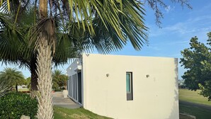 A modern building with a small window, surrounded by grass and palm trees. - Blue Skies Large Studio Apartment (Aguadilla)