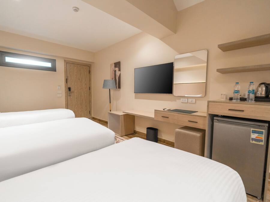 Habitación de hotel con dos camas, un escritorio con silla, televisor, espejo, refrigerador pequeño y un cubo de basura.