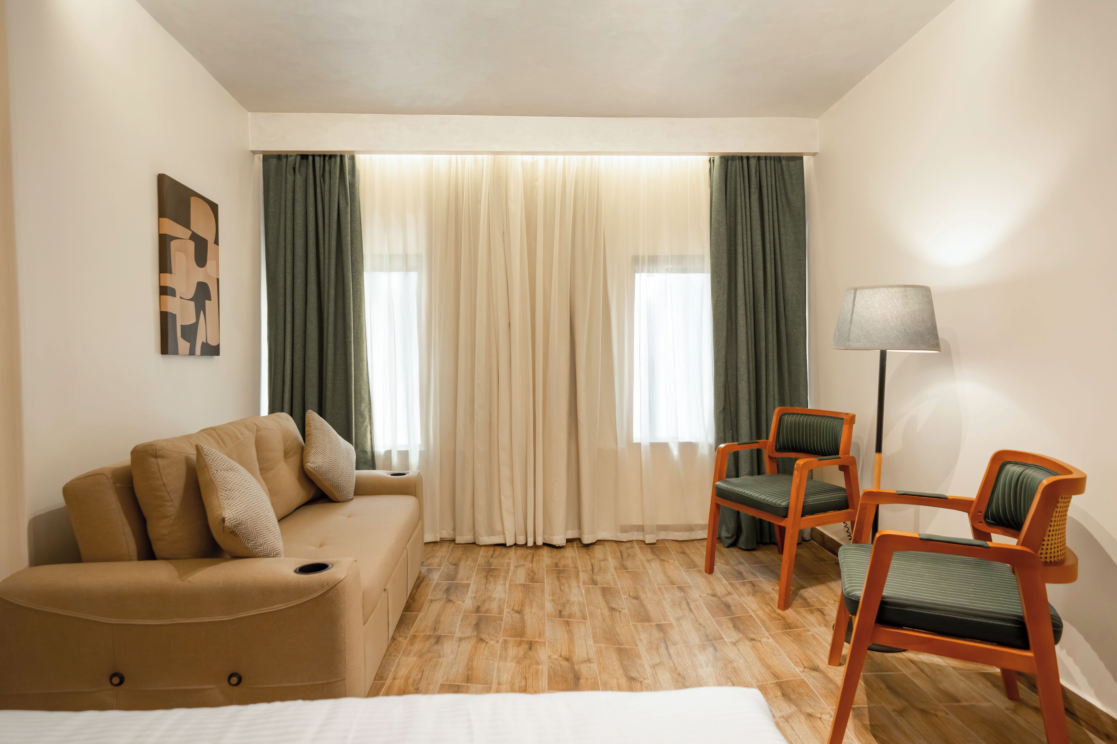 A hotel room with a beige sofa, two green chairs, a lamp, and a painting on the wall.