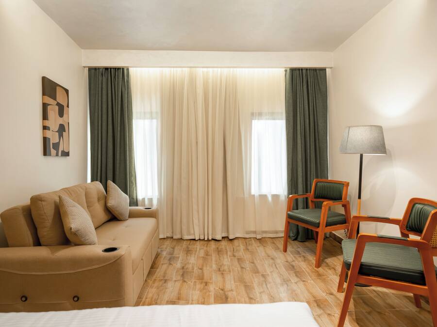 Habitación de hotel con un sofá color beige, dos sillones verdes, una lámpara y un cuadro en la pared.