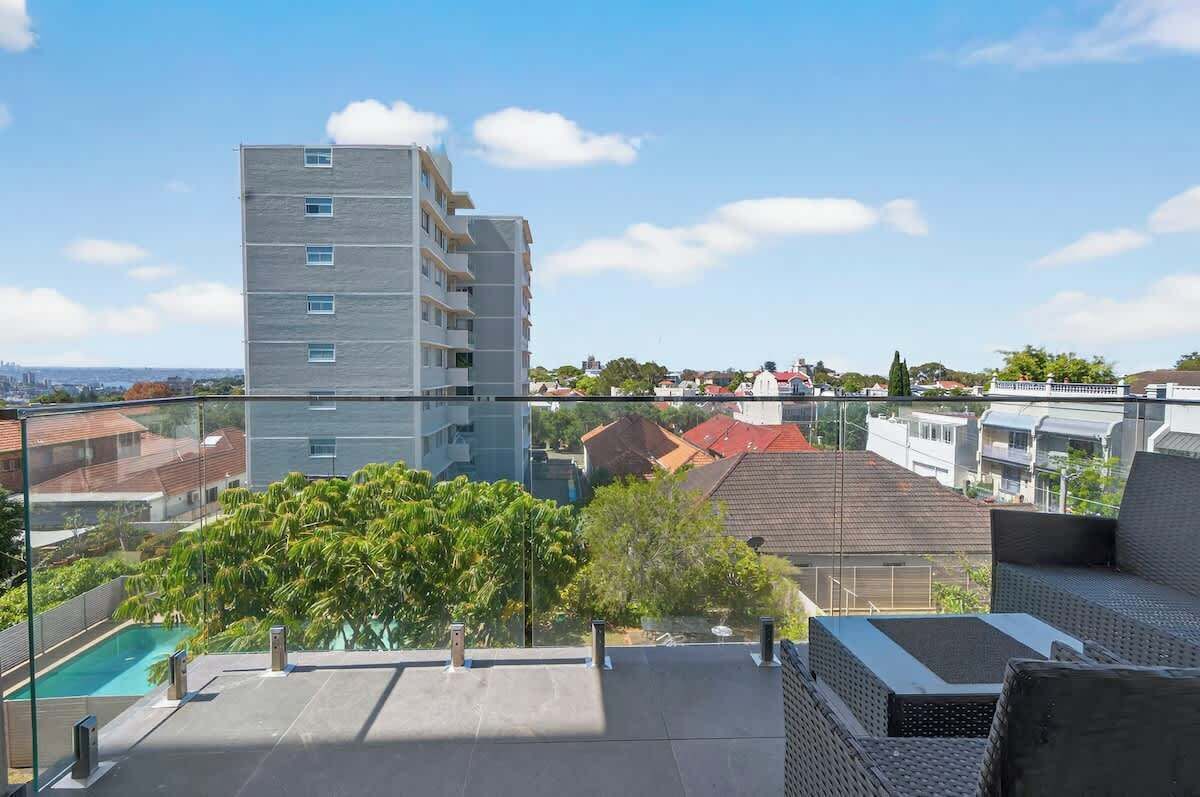 Un edificio moderno con piscina en la azotea y zona de estar al aire libre, rodeado de vegetación y otros edificios residenciales.