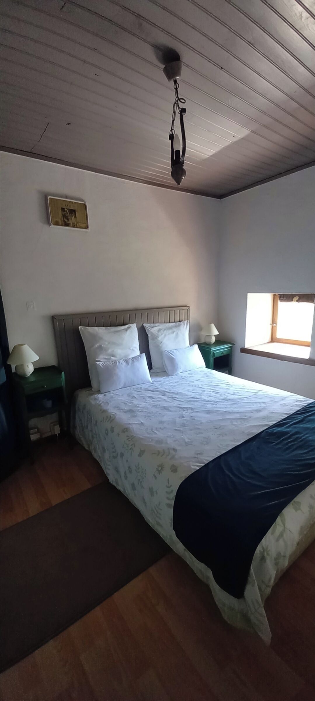 A bedroom with a large bed, a wooden floor, a ceiling fan, a nightstand with a lamp, and a window with natural light.