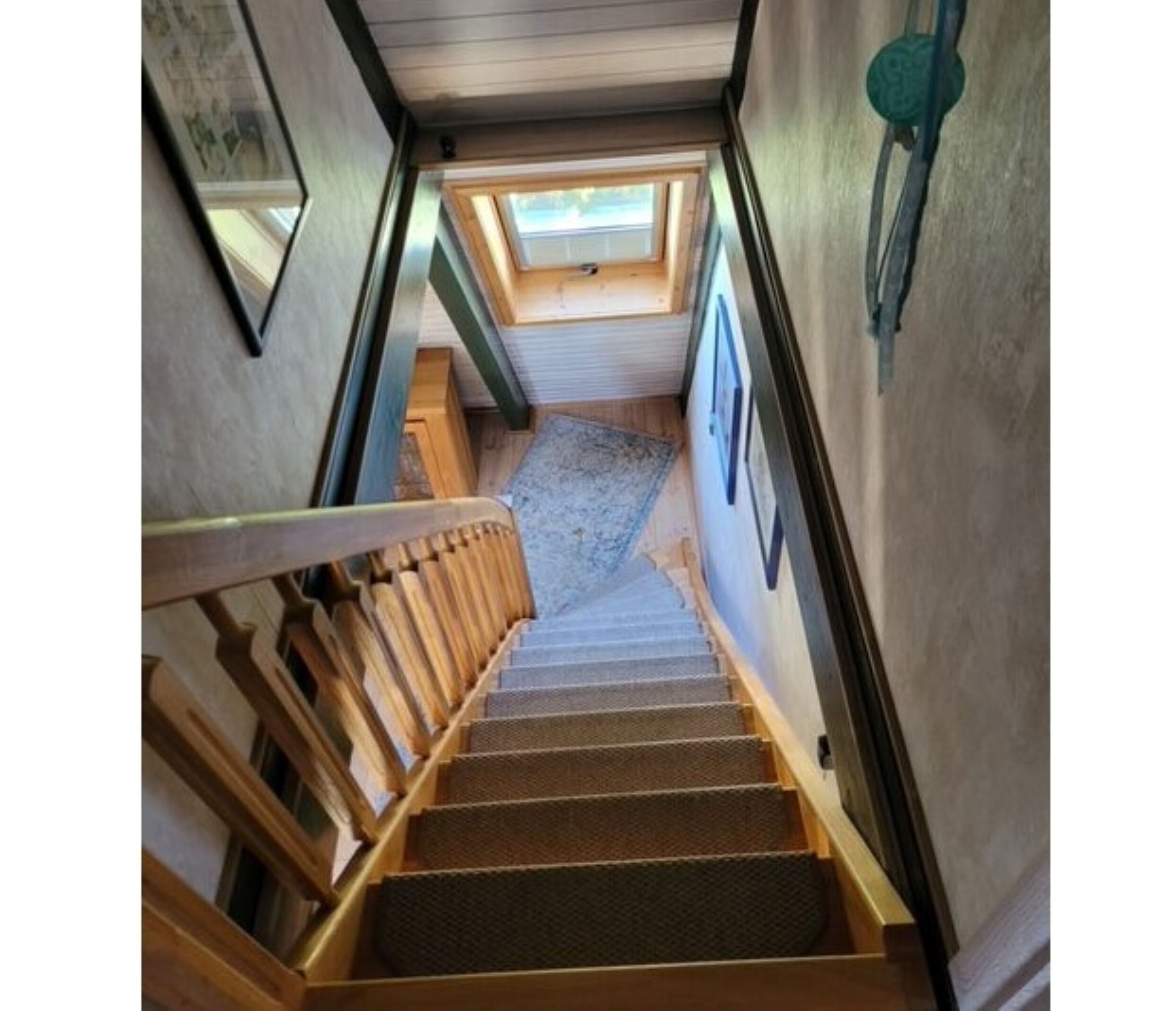 Un escalier étroit avec des rampes en bois et un sol recouvert de moquette, menant à une pièce dotée d’une lucarne.