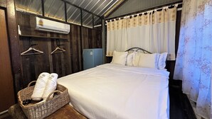 A single bed with white linens, a small blue refrigerator, a wicker basket with towels, and a wooden bench. - Cashew Garden House (Tambon Chalong)
