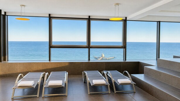 A modern indoor pool area with large windows overlooking the sea, sun loungers, and a clear view of a pier. - Exclusive beach shell vacation apartment with sauna and pool. (Sellin)