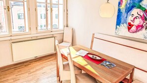 Un coin repas avec une table en bois, des chaises blanches, un radiateur et une fenêtre à carreaux.