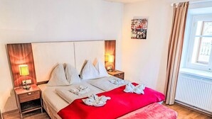 Una habitación de hotel con una cama grande, dos lámparas de noche, una manta roja y un ventanal con cortinas.