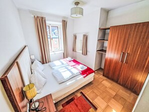 Un dormitorio con cama, armario de madera, ventana con cortinas, lámpara de noche y radiador.