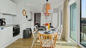 Een moderne keuken met witte kastjes, een eethoek met een houten tafel en stoelen, een hangende hanglamp en een groot raam met gordijnen.