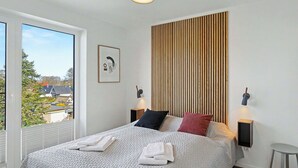 Een moderne slaapkamer met een groot bed, twee bedlampen, een wand van houten latten, een ingelijst kunstwerk en uitzicht door een groot raam.