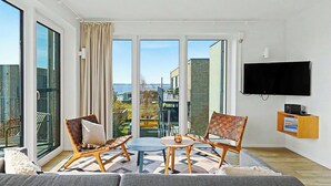 Een moderne woonkamer met een bank, twee rieten stoelen, een kleine tafel en een aan de muur bevestigde televisie.