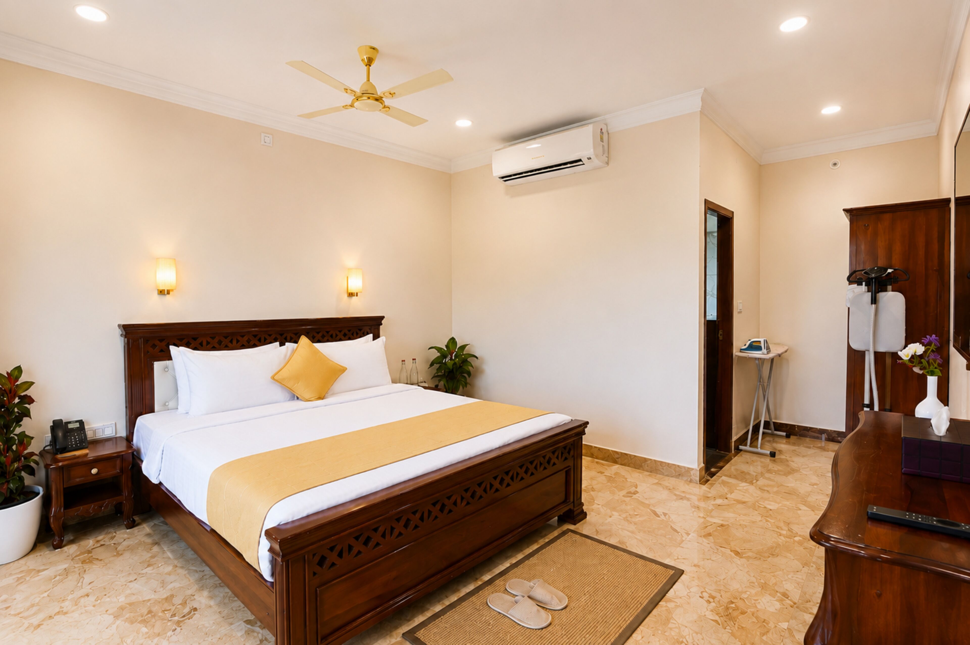 A hotel room with a wooden bed, a ceiling fan, an air conditioning unit, a bedside table with a phone, a potted plant, and a bathroom with a mirror and a towel rack.