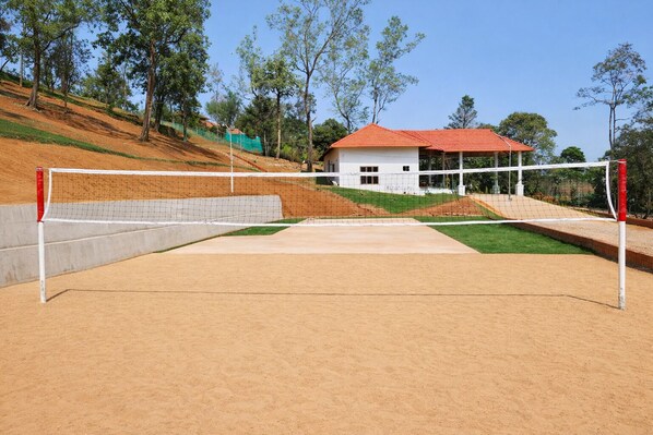 Un terrain de volley-ball avec un filet, un petit bâtiment doté d’un toit rouge et, au fond, une colline couverte d’herbe.