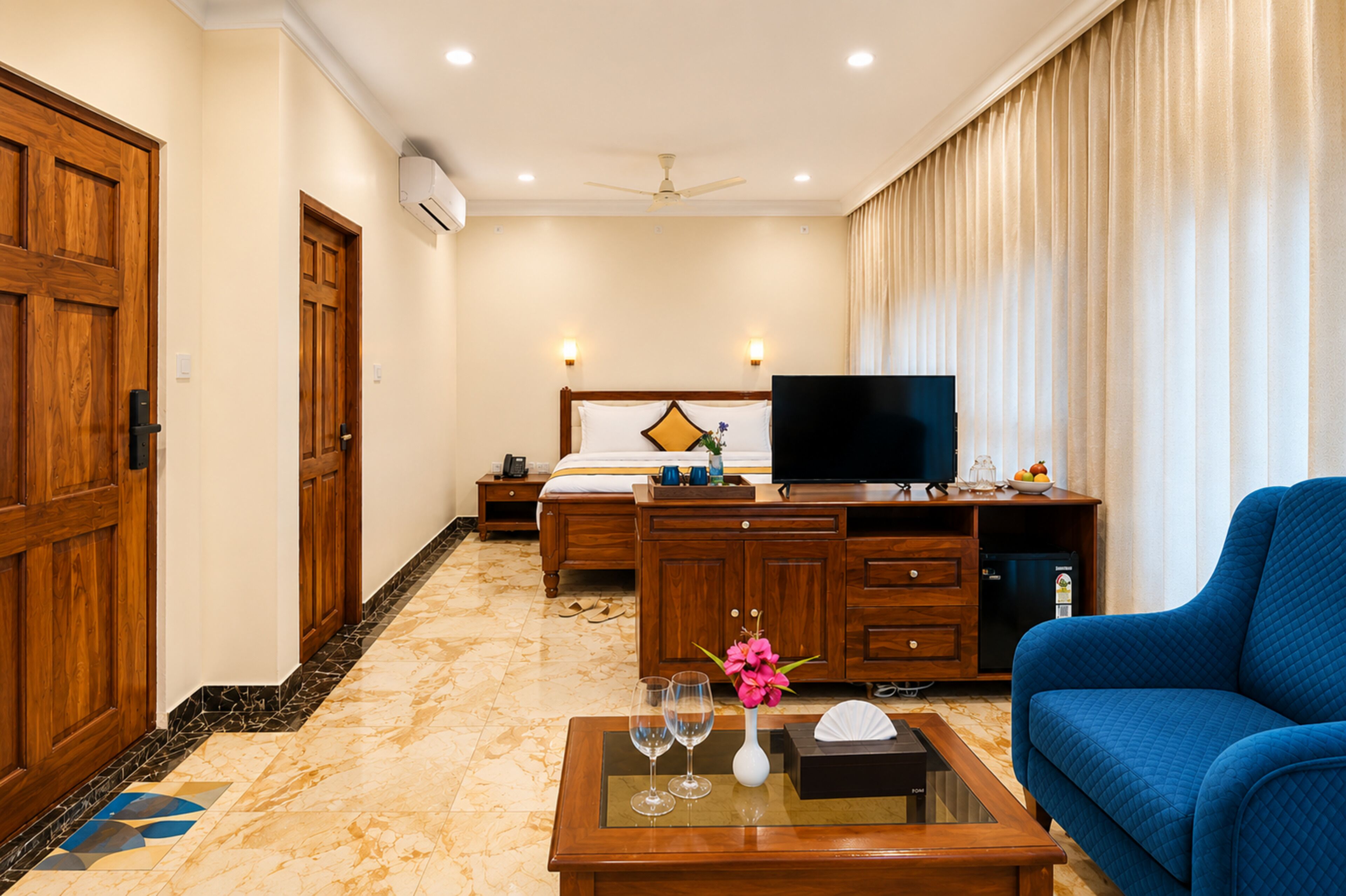 A hotel room with a bed, a desk with a flat-screen TV, a blue sofa, and a coffee table with a vase and wine glass.