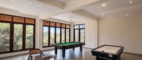 Una habitación amplia con mesa de billar, mesa de futbolín y mesa de ajedrez. Amplias ventanas ofrecen luz natural y vistas a la vegetación exterior.