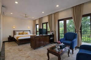 A hotel room with a bed, a wooden dresser, a blue armchair, and a balcony with a view of trees. - Astralis Resort Coorg (Madikeri)