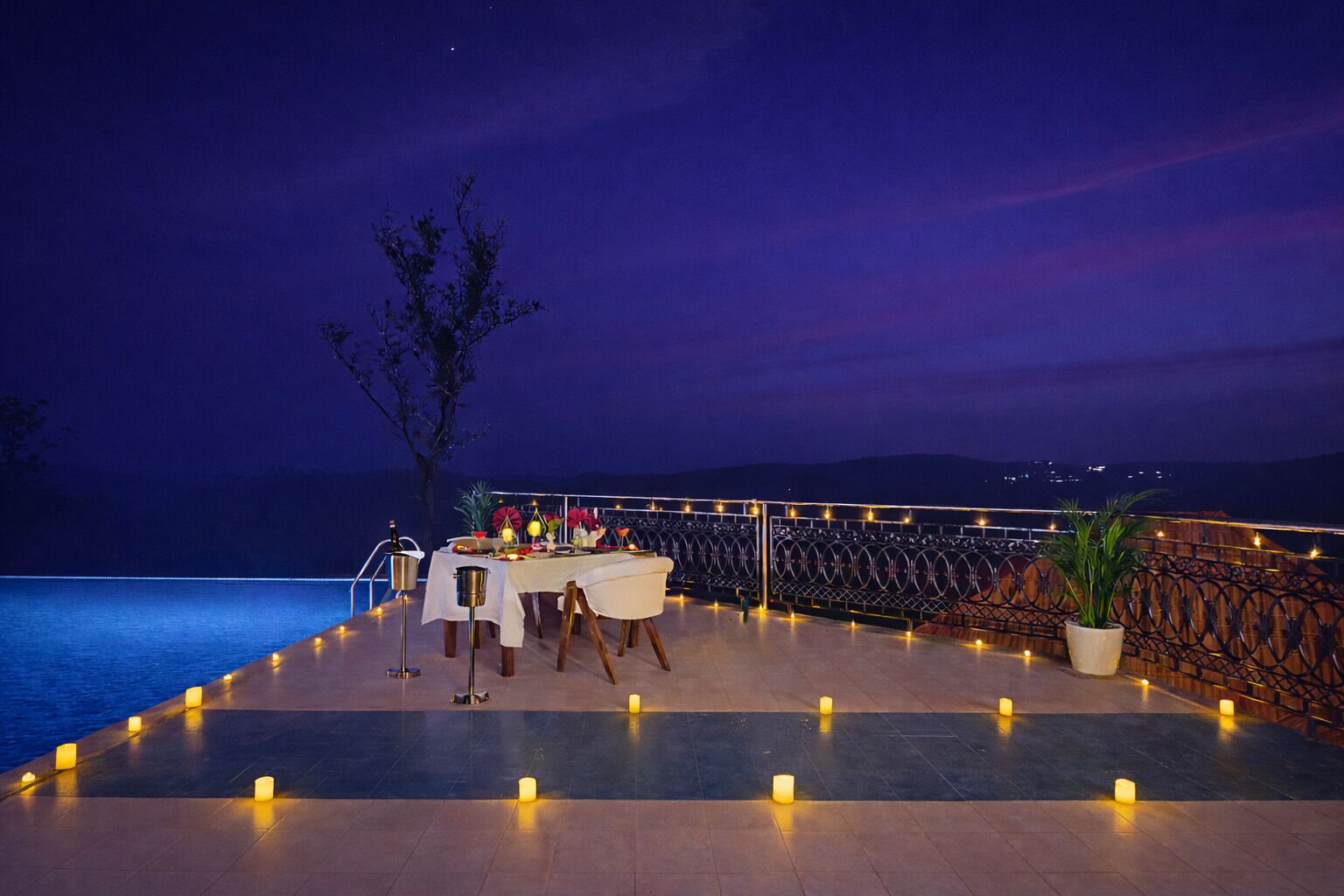 Un área de comedor al aire libre con una mesa dispuesta para dos personas, rodeada de velas, con vista a un cuerpo de agua y un paisaje montañoso.