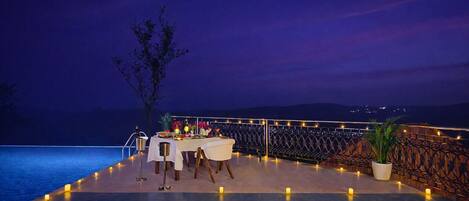 Un área de comedor al aire libre con una mesa dispuesta para dos personas, rodeada de velas, con vista a un cuerpo de agua y un paisaje montañoso.