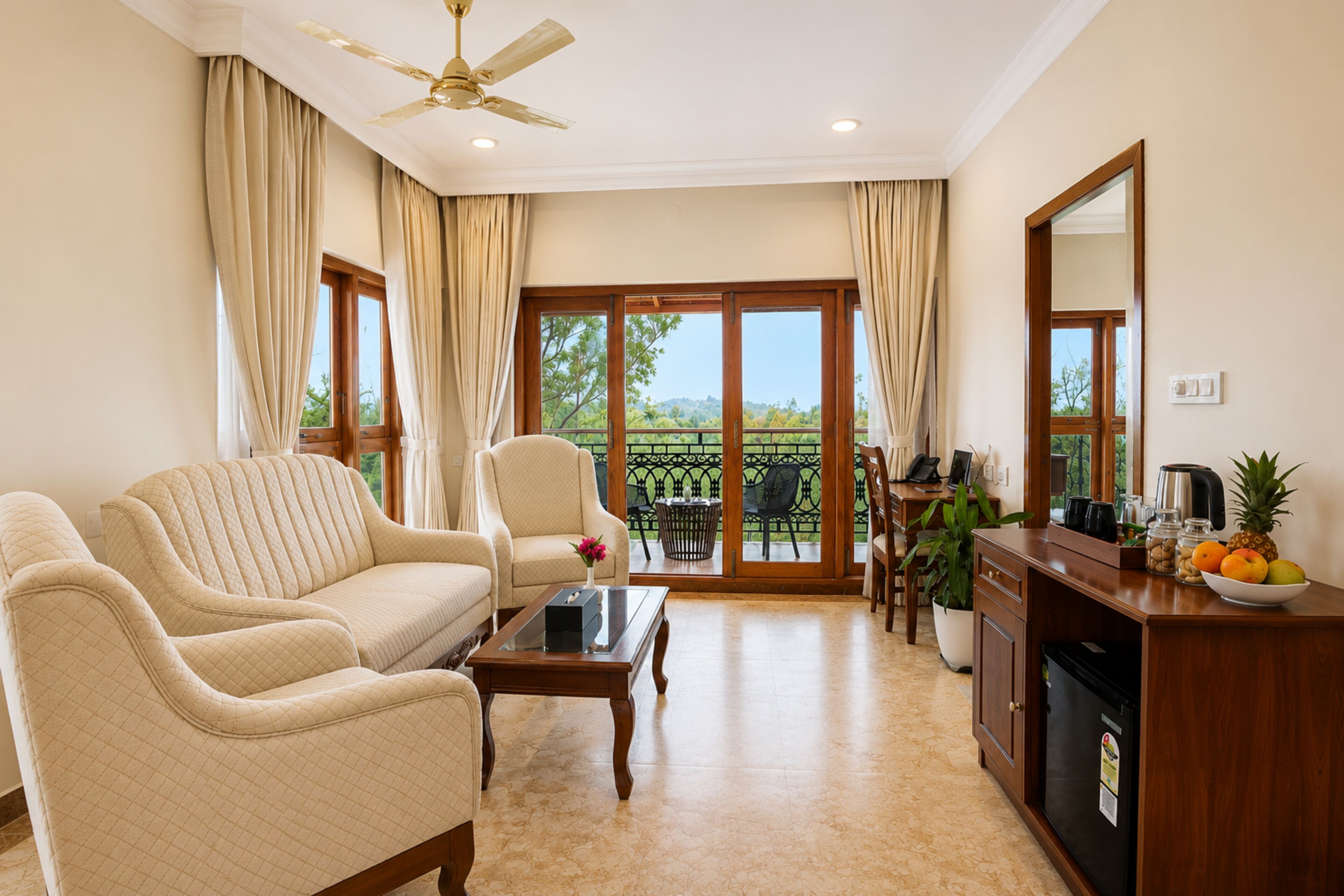 A living room with a sofa, armchair, coffee table, and a dining area with a table and chairs. There is a kitchen area with a refrigerator, coffee maker, and fruit bowl. The room has large windows with curtains, a ceiling fan, and a view of greenery outside.