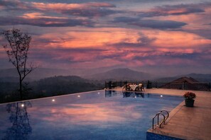 An infinity pool with a dining area and a view of mountains during sunset. - Astralis Resort Coorg (Madikeri)