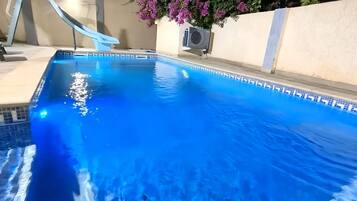 Une piscine avec une eau d’un bleu limpide, un toboggan et une plante en pot ornée de fleurs violettes.