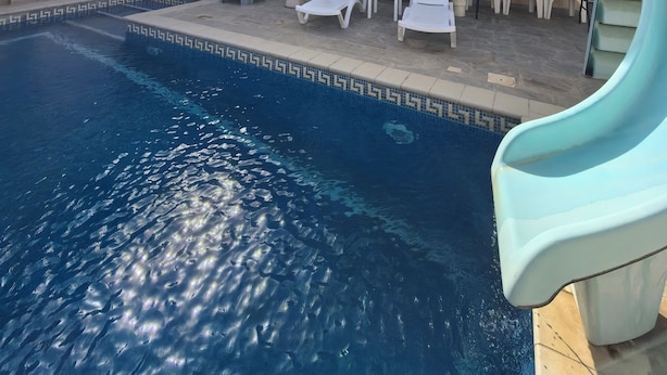 En swimmingpool med solstolar och en vattenrutschkana, omgiven av en byggnad med ett tak täckt av tegelpannor och väggar med ett dekorativt mönster.