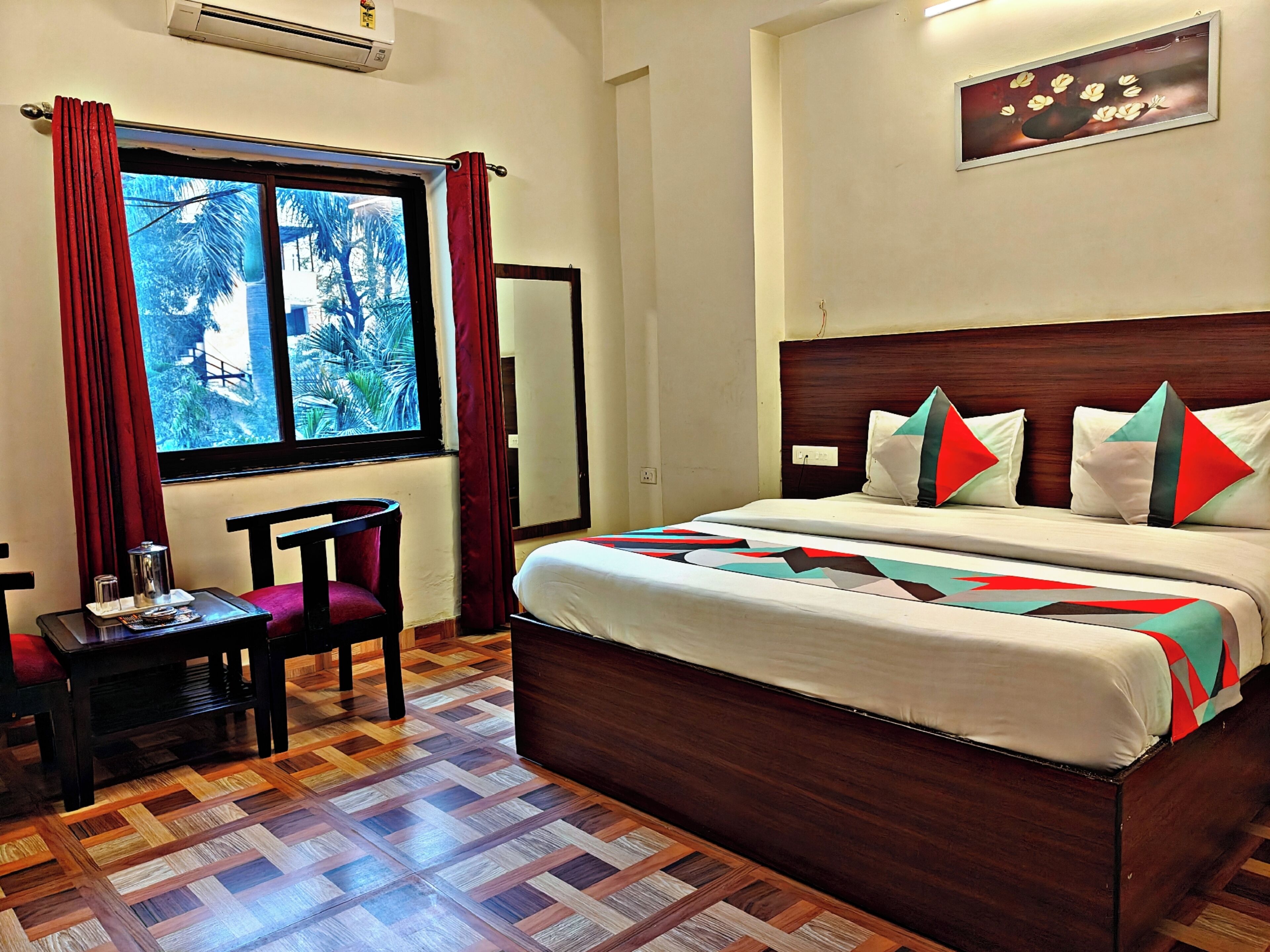 A hotel room with a bed, a chair, a small table, a window with curtains, and a wall-mounted mirror.