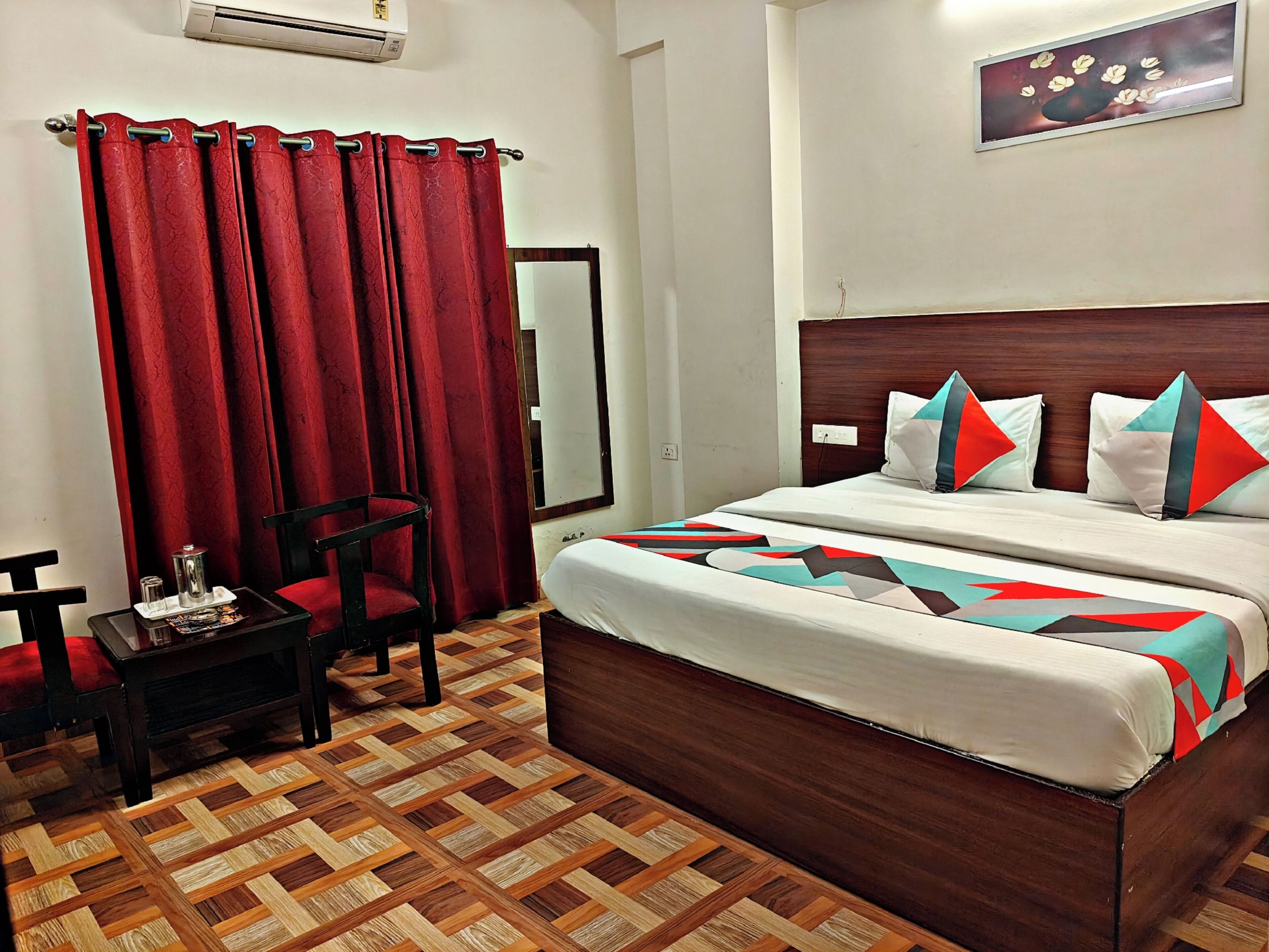 A hotel room with a large bed, a wooden headboard, a red curtain, a small table with a cup and saucer, and a patterned floor.