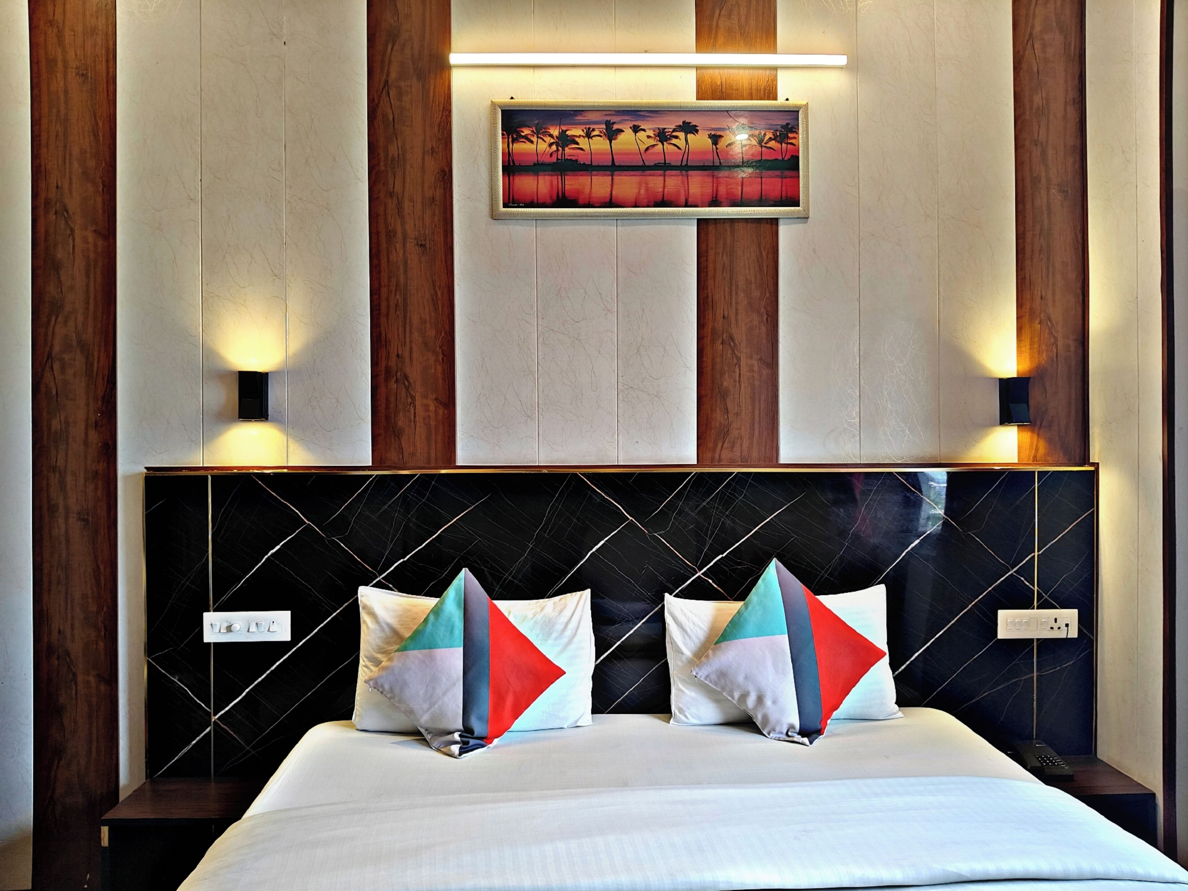 A hotel room with a bed, two wall-mounted lights, a painting, and a power outlet.