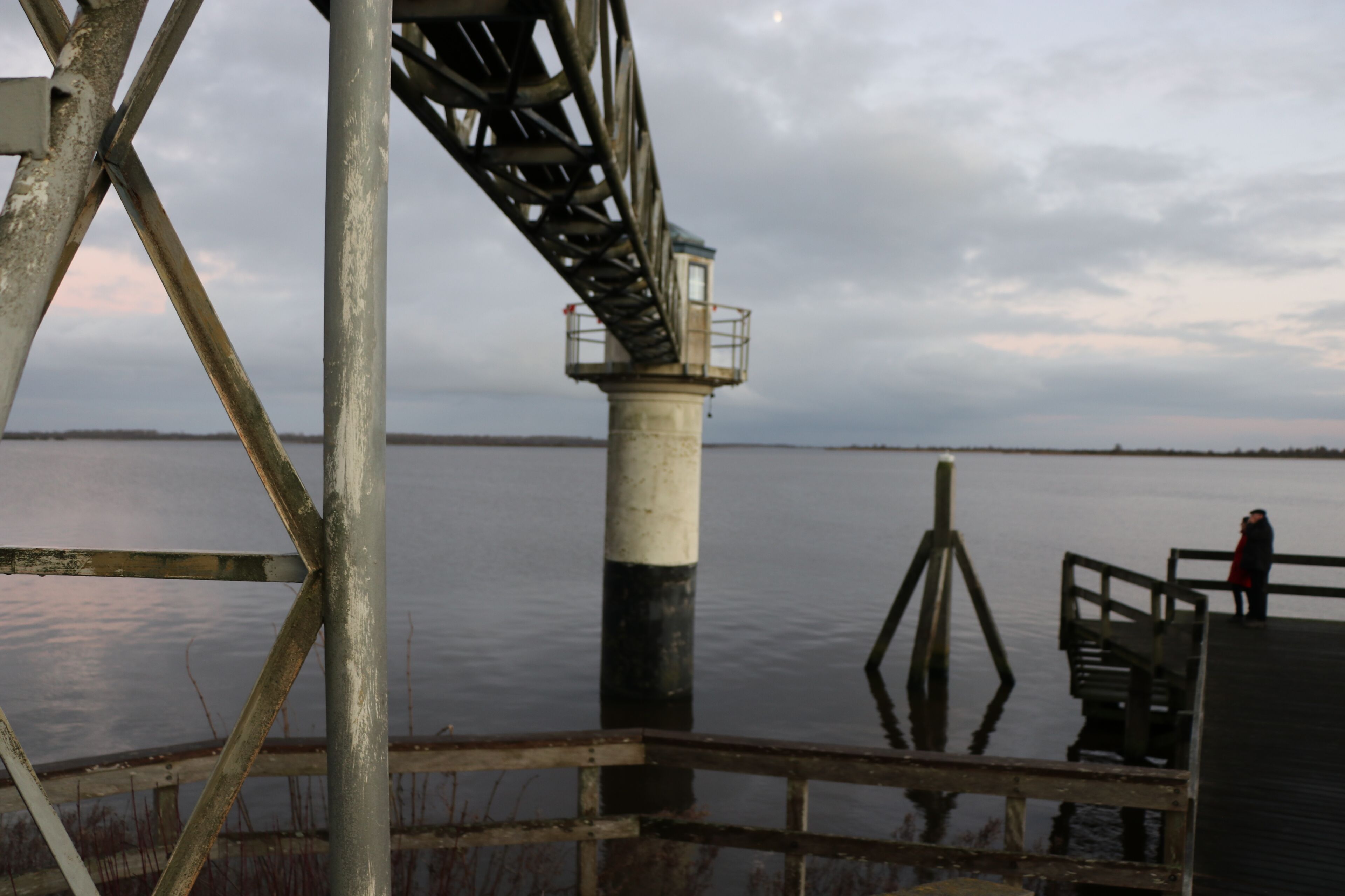 Una estructura de faro con armazón metálico, un muelle de madera y una masa de agua tranquila al fondo.