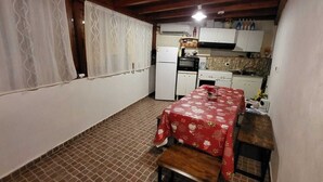 Een kleine keuken met een koelkast, oven en spoelbak. Er staat een tafel met een rode tafelkleed en een bankje. De vloer is betegeld en er is een raam met gordijnen.