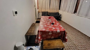 Een kamer met een lange tafel, afgedekt met een rode tafelkleed met bloemen, een bank en een klein krukje.