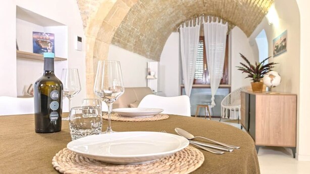 A dining table set with plates, cutlery, and wine glasses, with a bottle of wine and a plant in the background. - Il Viandante, Loft Indipendente nel centro di Cagliari (Cagliari)