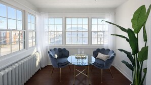 A room with a round glass table, two blue armchairs, a large green plant, and a view of buildings through large windows. - Pet Friendly Estate, Sleeps 20 Includes Linens, Towels, Beach Tags & Parking (Ocean City)