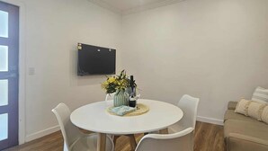 Un espace repas moderne avec une table ronde, quatre chaises blanches et un téléviseur à écran plat fixé au mur.