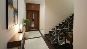 A modern hallway with a wooden door, a staircase, a desk with a plant, and a framed artwork on the wall. - Hotel Anmol One  (Indore)