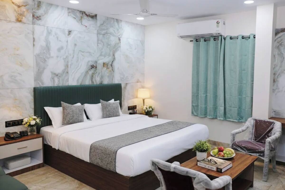 Una habitación de hotel moderna con pared de mármol, cabecero tapizado en verde, mesita de noche de madera, una lámpara en la mesita, cortina verde, una silla y una pequeña mesa con frutas y un libro.