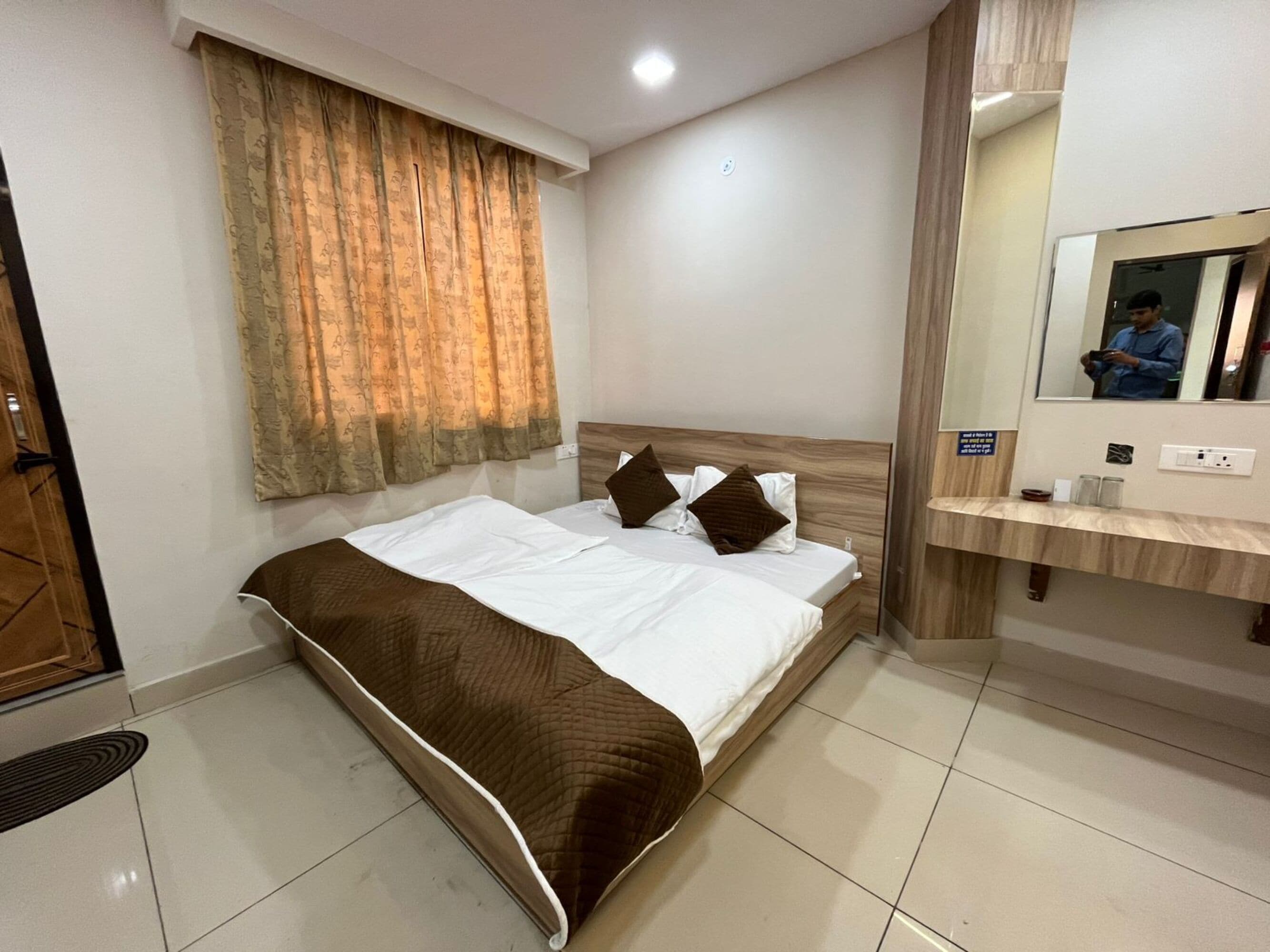 Quarto de hotel com cama, cabeceira de madeira, espelho, penteadeira com bancada e janela com cortinas.