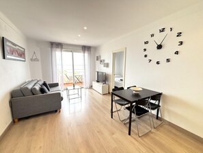 A modern living room with a sofa, dining table, chairs, a TV, and a wall clock. - Renovated beachfront apartment with AC and balcony in Roses (Roses)