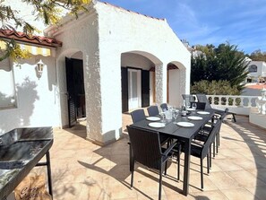 An outdoor patio with a table set for dining, a grill, and a white building with arches. - Charming Private House with Garden, 300m from Canyelles Beach - Sleeps 6, Roses, Spain (Roses)
