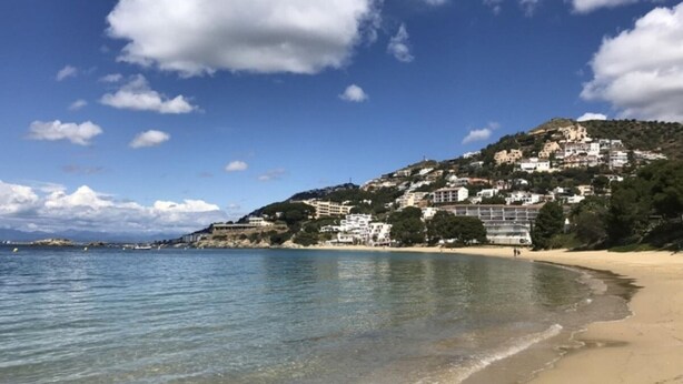 Una playa costera con aguas cristalinas, un litoral arenoso y edificios en una ladera.