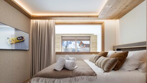 Hotellihuone, jossa on sänky, ikkuna, josta avautuu lumihuippuisten vuorten maisema, seinälle asennettu taulutelevisio ja puinen katto.