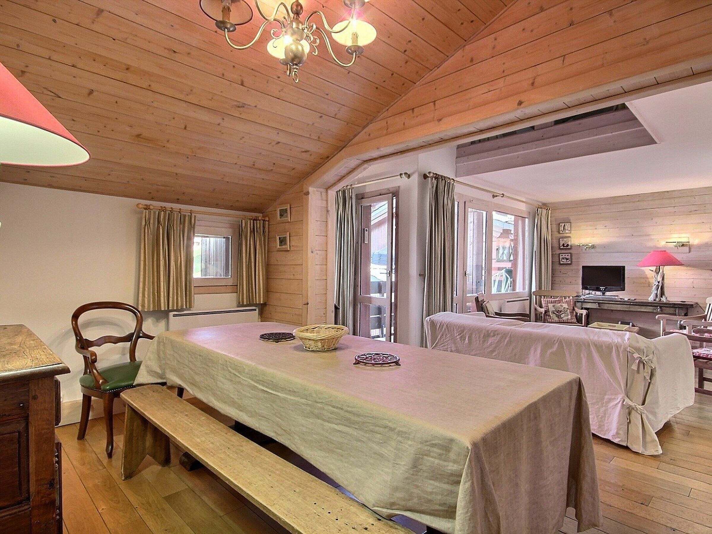 A cozy room with wooden walls and a high ceiling, featuring a dining table with a bench, a television, and a window with curtains.