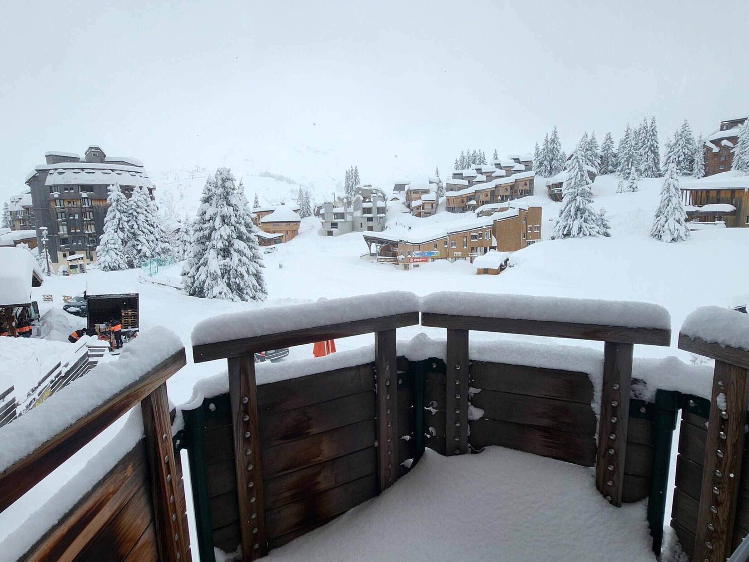 A snow-covered balcony with wooden railings overlooking a residential area with houses and trees.