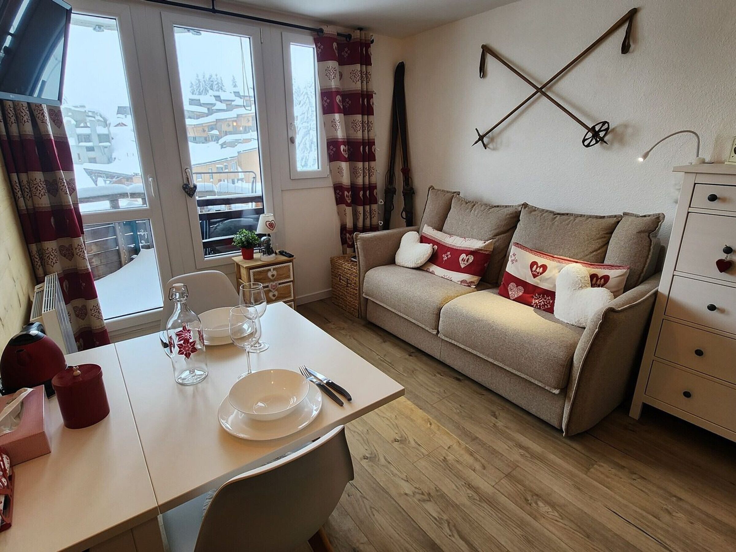Un salon chaleureux avec un canapé, une table à manger dressée avec des assiettes et des verres, une petite table d’appoint avec une lampe, et une vue sur des bâtiments enneigés par la fenêtre.