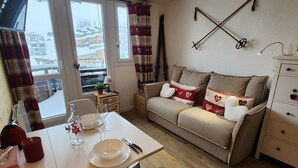 Una sala acogedora con sofá, una mesa de comedor dispuesta con platos y vasos, una mesita auxiliar con lámpara, y vistas de edificios nevados a través de la ventana.