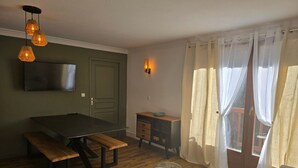 Ein Zimmer mit Holzboden, einem Esstisch mit Bänken, einem an der Wand montierten Fernseher, einem kleinen weißen Hocker und einem Fenster mit durchscheinenden Vorhängen.